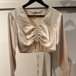 Beige satinblus från NA-KD - Elegant beige blus i satin från NA-KD med v-ringning och långa ärmar. Blusen har en snygg snörning framtill som ger en fin detalj och är croppad för en modern look.