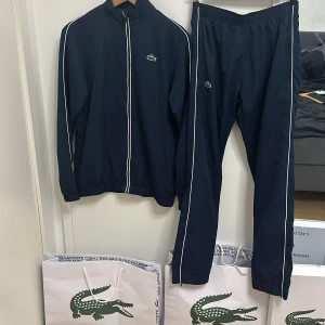 Mörkblå Lacoste tracksuit  - Snyggt mörkblått träningsset från Lacoste med vita detaljer, den är på 10/10 skick helt ny. Jackan har dragkedja och broderad logga på bröstet samt en stor logga på ryggen. Byxorna har elastisk midja och matchande logga. Perfekt för en sportig look! Org pris 2900 mitt pris 1499 priset går att diskuteras, storleken är xs