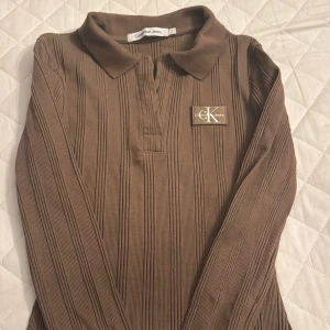 Brun ribbad klänning från Calvin Klein Jeans - Snygg brun ribbad klänning från Calvin Klein Jeans med långärmad design och klassisk krage. Perfekt för en stilren look med diskret logga på bröstet.