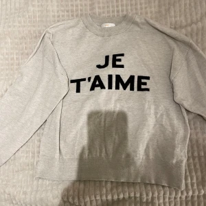 Grå tröja från Gina Tricot med text - Säljer en grå tröja från Gina Tricot med texten 'JE T'AIME' i svart på framsidan. Tröjan har långa ärmar och en rund halsringning. 