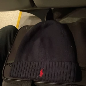 Mörkblå mössa från Ralph Lauren - Snygg mörkblå mössa från Ralph Lauren med röd broderad logga framtill. Den är ribbad vid kanten och perfekt för kyliga dagar.