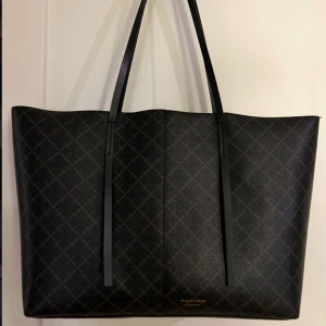 Svart shoppingväska från By Malene Birger - Snygg svart shoppingväska från By Malene Birger med ett diskret rutigt mönster. Väskan har långa handtag och är perfekt för att bära med sig allt man behöver. Tillverkad i ett slitstarkt material med stilren design. Ett av handtagen är något nött men utöver det så är väskan i väldigt bra skick. (Ordinarie pris 2795kr)