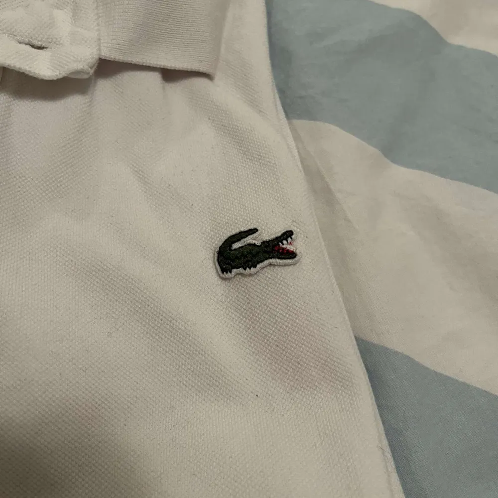 Säljer en klassisk vit pikétröja från Lacoste med korta ärmar och den ikoniska krokodilloggan på bröstet. Tröjan har en krage med knappar.. T-paidat.