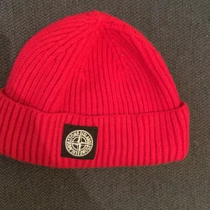 Röd stickad mössa från Stone Island - Snygg röd stickad mössa från Stone Island med ribbad design och logotyp framtill. Perfekt för att hålla värmen under kyliga dagar.