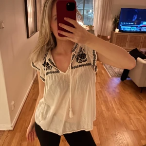 Vit blus med broderi från H&M - Säljer en vit blus från H&M med svart broderi och knytband framtill. Blusen har korta ärmar och en avslappnad passform, perfekt för en bohemisk look.