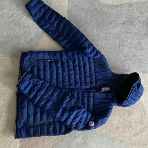 Äkta Moncler jacka - Säljer en snygg blå quiltad jacka från Moncler. Jackan har en dragkedja framtill och två praktiska fickor med dragkedjor. Perfekt för kyligare dagar med sin stilrena design och varma material.