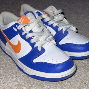 Nike Dunk sneakers i blått och vitt - Snygga Nike Dunk sneakers i blått och vitt med orange detaljer. Skorna har klassisk snörning och en sportig design som passar perfekt för en avslappnad stil. Helt nya köpta för 1100kr och säljer för 450kr