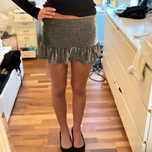 Glittrig kortkjol med volang - Snygg glittrig kortkjol i silver med volangdetalj. Perfekt för en kväll ute eller en festlig tillställning. Kjolen har en tight passform som framhäver figuren.