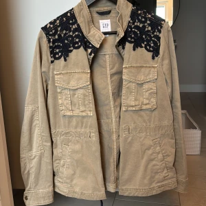 Beige jacka med spetsdetaljer från GAP - Snygg beige jacka från GAP med unika svarta spetsdetaljer på axlarna. Jackan har flera praktiska fickor framtill och knäppning med knappar. Perfekt för en stilren look.