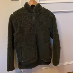 Grön teddyjacka med dragkedja - Säljer en mysig grön teddyjacka med dragkedja framtill. Jackan är köpt på j Lindberg för ungefär ett år sedan och är använd sparsamt och i fantastisk skick