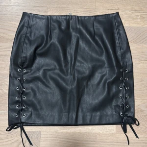 Svart kjol med snörning från Rebecca Stella - Snygg svart kjol i skinnimitation från Rebecca Stella. Kjolen har en tight passform med snörningsdetaljer på sidorna och en dragkedja bak.