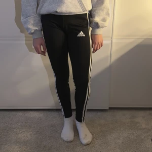 Svarta leggings från Adidas - Snygga svarta leggings från Adidas med de klassiska tre vita ränderna längs sidorna. Perfekta för träning eller en avslappnad dag. De har en tight passform och är tillverkade i ett stretchigt material för bästa komfort.