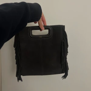 Mörk grå mockaväska med fransar  - Snygg mörk grå väska i mocka med fransar på sidorna och ett praktiskt handtag. Väskan har även en avtagbar axelrem för extra flexibilitet.