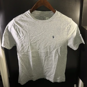 Grå t-shirt från Ralph Lauren - Säljer en klassisk grå t-shirt från Ralph Lauren med korta ärmar och en liten broderad logga på bröstet. Perfekt för en avslappnad stil. Hör av er vid minsta fundering🙌