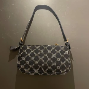 Svart axelväska med mönster - Snygg svart axelväska med ett elegant cirkelmönster i vitt. Väskan har en praktisk dragkedja och guldfärgade detaljer vid remfästena. Perfekt för att ge en stilren touch till din outfit.