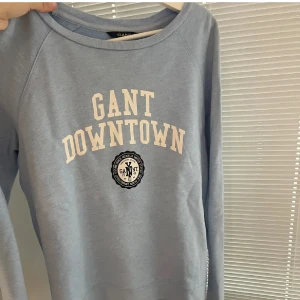 Blå sweatshirt från Gant - Säljer en snygg blå sweatshirt från Gant med texten 'GANT DOWNTOWN' i vitt på framsidan. Tröjan har långa ärmar och en rund halsringning. Perfekt för en avslappnad stil.