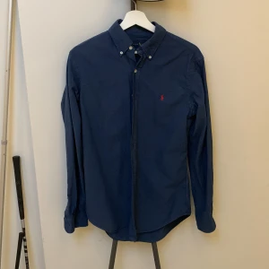 Blå skjorta från Ralph Lauren - Snygg blå skjorta från Ralph Lauren i slim fit. Skjortan har långa ärmar och knappar framtill. Den är broderad med Ralph Laurens ikoniska röda logotyp på bröstet.ny pris 1500kr mitt pris 699kr. Skick 9/10