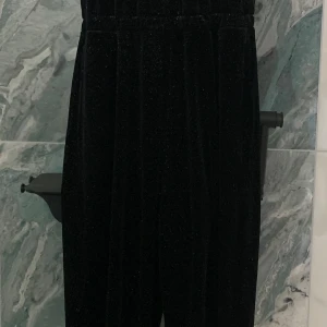 Svart glittrig byxdress från Lindex - Snygg svart byxdress från Lindex med glittrigt material. Perfekt för en festlig look. Byxdressen har korta ärmar med volangdetaljer och en bekväm passform.