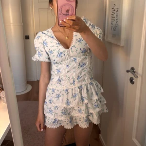 Blommig klänning med volanger - Charmig kortklänning med blommigt mönster i blått och grönt på vit botten. Klänningen har en v-ringad hals och korta ärmar med volangdetaljer. Dragkedja på sidan, ej använd och prislapp sitter kvar. Passar som både XS och XXS.