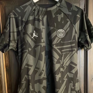 Svart PSG fotbollströja från Jordan - Snygg svart PSG fotbollströja från Jordan med ett coolt kamouflagemönster. Tröjan har helt oanvänd och passar alla.