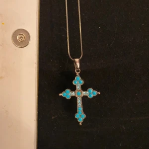 Halsband med korshänge i turkos och silver - Halsband med ett korshänge dekorerat med turkosa stenar och silverdetaljer. Perfekt för att ge en färgklick till din outfit.