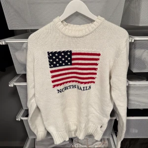 Vit stickad tröja från North Sails - Säljer en vit stickad tröja från North Sails med en stor amerikansk flagga i rött och blått på framsidan. Tröjan har långa ärmar och ribbstickade muddar vid ärmslut och nederkant. Perfekt för kyliga dagar! Storlek L men är lite liten så L/M