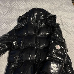 Svart Maya jacka från Moncler - Snygg svart Maya jacka  från Moncler med glansig finish och dragkedja framtill. Jackan har en hög krage och en ikonisk Moncler-logga på ärmen. Perfekt för kalla dagar med sin vadderade design.Säljer  på grund av den  är för stor det är M 
