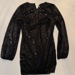 Svart glittrig klänning - Snygg svart klänning med glittriga detaljer som ger en festlig look. Klänningen har långa ärmar och en kort längd, perfekt för en utekväll. Den är tillverkad i ett material som ger en skimrande effekt.
