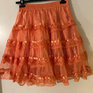 Orange tyllkjol med spetsdetaljer - Charmig orange tyllkjol med flera lager och spetsdetaljer. Kjolen har en elastisk midja för bekväm passform och volanger som ger extra volym. Perfekt för att lägga till en färgglad touch till din outfit.