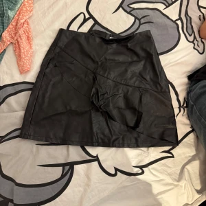 Svart skinnkjol - Skitsnygg svart skinnkjol. Kjolen har en dragkedja baktill och en asymmetrisk detalj framtill som ger en modern touch. Perfekt för att ge din outfit en edgy look. Lite skrynlig men aldrig använd. Osäker på storlek men tror den hade passat XS/S