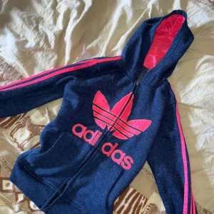 Adidas kofta med neonrosa detaljer - Snygg blå huvtröja från Adidas med neon rosa detaljer och dragkedja. Tröjan har en stor logga på framsidan och rosa ränder längs ärmarna. Använd men i fint skick