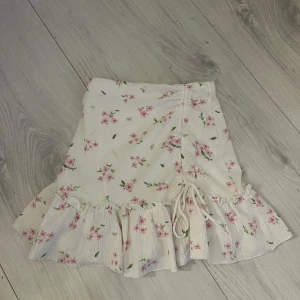 Blommig kjol med volang - Charmig blommig kjol i ljusa färger med volangdetaljer och knytning framtill. Perfekt för en somrig look.