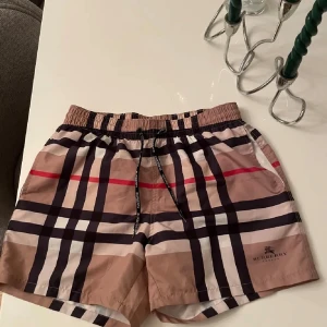 Rutiga badshorts från Burberry - Snygga badshorts från Burberry med klassiskt rutigt mönster i beige, svart, vit och röd. De har en elastisk midja med snörning för perfekt passform. Perfekta för sommarens alla badäventyr! Säljer dessa pågrund av att dem va lite för små för mig. Kan såklart gå ner i pris vid snabb affär, hör av er vid frågor!