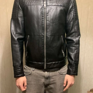 Svart skinnjacka från Jack & Jones - Snygg svart skinnjacka från Jack & Jones med dragkedja framtill. Jackan har quiltade detaljer på axlar och ärmar samt ribbade muddar vid ärmslut och nederkant. Perfekt för en stilren look.