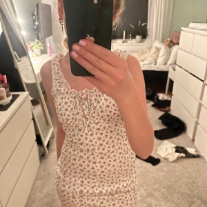 Blommig klänning med volangdetaljer - Söt blommig klänning i vitt med små röda blommor. Den har volangdetaljer vid axelbanden och en knytning framtill. Perfekt för en somrig look!