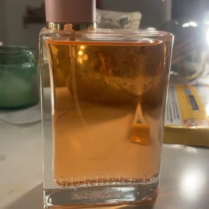 Parfym från Burberry - Elegant parfym från Burberry i en stilren glasflaska med rosa lock och guldiga detaljer. Doften har en varm och inbjudande ton, perfekt för den som vill ha en sofistikerad touch. Flaskan är genomskinlig och visar den ljusrosa vätskan inuti. Har kanske använt den 2 gånger 