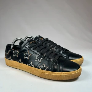 Saint Laurent skor - Snygga svarta sneakers från Saint Laurent med stjärnmönster i silver. Skorna har en klassisk design med snörning och en bekväm gummisula i beige. Perfekta för en stilren look.