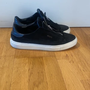 Axel Arigato  - Snygga svarta sneakers från Arigato med dragkedja framtill. Skorna har en stilren design med vit sula och är tillverkade i mocka! Nypris ca 2000kr. Box och påse ingår! Strl 42, passar 43. Skick 7/10