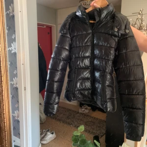 Svart populär glansig jacka från 157 - Snygg svart dunjacka från 157 i glansigt material. Jackan har en dragkedja framtill och en praktisk huva. Perfekt för kyliga dagar med sin vadderade design som håller dig varm och bekväm. Storlek xxs men funkar även på mig som är xs/s