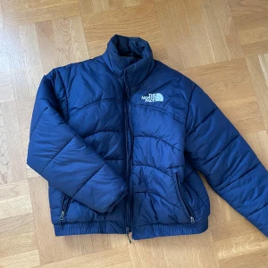 Blå dunjacka från The North Face - Säljer en blå dunjacka från The North Face med dragkedja och hög krage. Jackan har långa ärmar och två fickor med dragkedja framtill. Perfekt för kyliga dagar. Den är köpt på zalando förra vintern