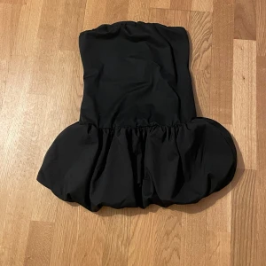 Svart axelbandslös klänning - Snygg svart axelbandslös klänning med volangdetalj vid nederkanten. Perfekt för en kväll ute eller en speciell tillställning. Klänningen har en elegant och stilren design som framhäver figuren. Aldrig använt! 