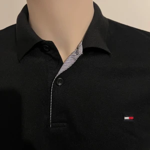 Svart pikétröja från Tommy Hilfiger - Snygg svart pikétröja från Tommy Hilfiger med klassisk krage och knappslå. Tröjan har en liten logotyp på bröstet och är perfekt för en stilren look. Tillverkad i ett bekvämt material som passar till många tillfällen.