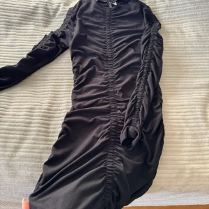Svart klänning från hm - Snygg svart klänning från hm med rynkade detaljer längs ärmarna och framsidan. Klänningen har långa ärmar och en figurnära passform som ger en elegant look. Perfekt för en kväll ute eller en speciell tillställning.