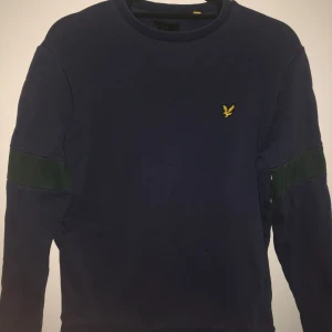 Mörkblå tröja från Lyle & Scott - Säljer en snygg mörkblå tröja från Lyle & Scott med deras ikoniska gula logga på bröstet. Tröjan har långa ärmar och en stilren design med gröna detaljer på ärmarna. Perfekt för en casual look!