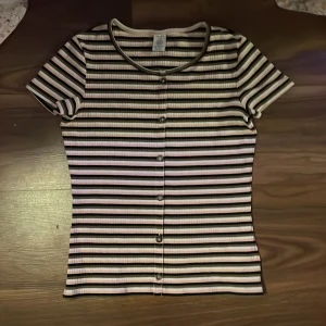  T-shirt - Säljer en t-shirt med knappar framtill Knapparna är endast för fint går inte att öppna! 💕 t-shirten kommer från Lindex och är i storlek 158/164 (12-14år). Den är randig och har ribbat tyg ⭐️