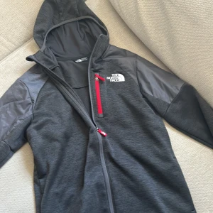 Grå jacka med huva från The North Face - Säljer en grå jacka med huva från The North Face. Jackan har en röd dragkedja på bröstfickan och en hel dragkedja framtill. Perfekt för kyligare dagar med sin stilrena design och praktiska fickor.
