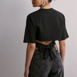 Svart kortärmad topp med knyt - Trendig svart kortärmad topp med knyt i ryggen. Perfekt för en stilren look med en twist. Toppen är croppad och har en modern design som passar till många olika outfits.