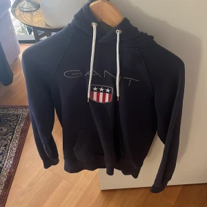 Mörkblå hoodie från GANT - Säljer en snygg mörkblå hoodie från GANT med ett stilrent tryck och logotyp på bröstet. Tröjan har en klassisk känguruficka och justerbar huva med vita snören. Perfekt för en avslappnad stil.