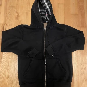 Svart hoodie från Burberry - Snygg svart hoodie från Burberry med klassiskt rutigt mönster i fodret. Den har en dragkedja framtill och en justerbar huva. Perfekt för en stilren look.