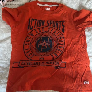 Orange t-shirt från WA - Säljer en cool orange t-shirt från WA med tryck 'Action Sports' och 'Surf Snow and Skateboarding'. Perfekt för en avslappnad stil. T-shirten har en rund hals och korta ärmar.
